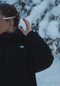 Black zip-up jacket with a hood, featuring a white New Balance logo, worn over a dark scarf, set against a snow-covered background.