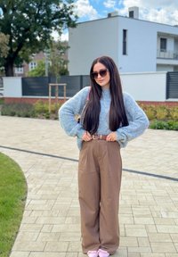 Overdimensioneret lyseblå strikket sweater parret med løse brune bukser. Langt brunt hår og solbriller, stående på en belagt sti i en have.