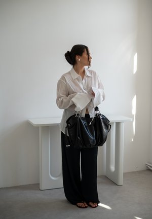 Mujer con camisa blanca y pantalones negros sosteniendo un gran bolso negro, de pie junto a una mesa blanca minimalista en una habitación luminosa con suelo de cemento.