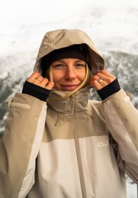 Beige vedenpitävä takki, jossa on huppu, vetoketjukiinnitys ja säädettävät hihansuut, puettuna ruskean neulotun pipo kanssa lumisessa maisemassa.