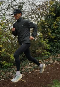 Casaco de corrida leve preto, leggings pretas, sapatos de corrida cinzentos com detalhes claros. Corredor em movimento com um boné e vários anéis.