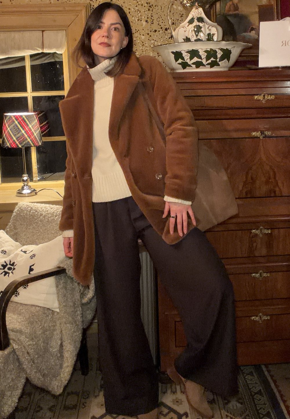 Manteau marron à double boutonnage en fourrure, porté sur un pull blanc côtelé. Pantalon large et sombre avec bottines beiges à la cheville. Texture douce, couleurs neutres.