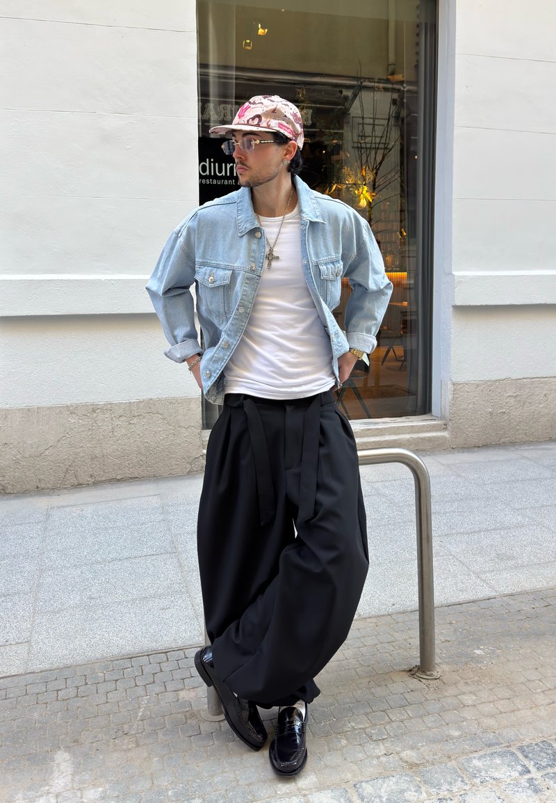 Ein Mann trägt eine hellblaue Jeansjacke, ein weißes Hemd, übergroße schwarze Hosen und eine rosa gemusterte Mütze und lehnt an einem Metallgeländer auf der Straße vor einem Gebäude.