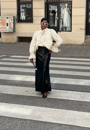 Mujer con blusa blanca de lunares, falda larga negra y gafas de sol blancas, posando con confianza en un paso de peatones de la ciudad.