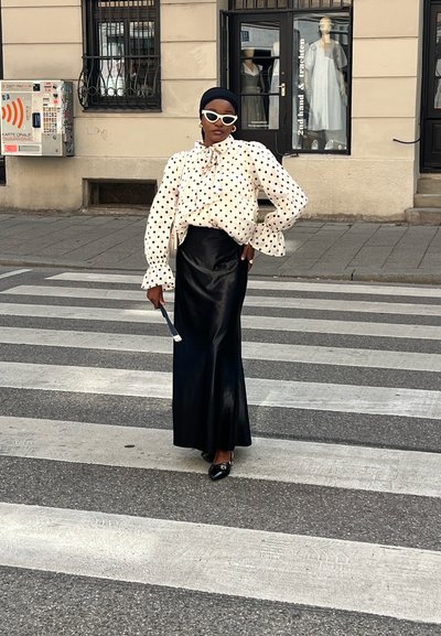 Mujer con blusa blanca de lunares, falda larga negra y gafas de sol blancas, posando con confianza en un paso de peatones de la ciudad.