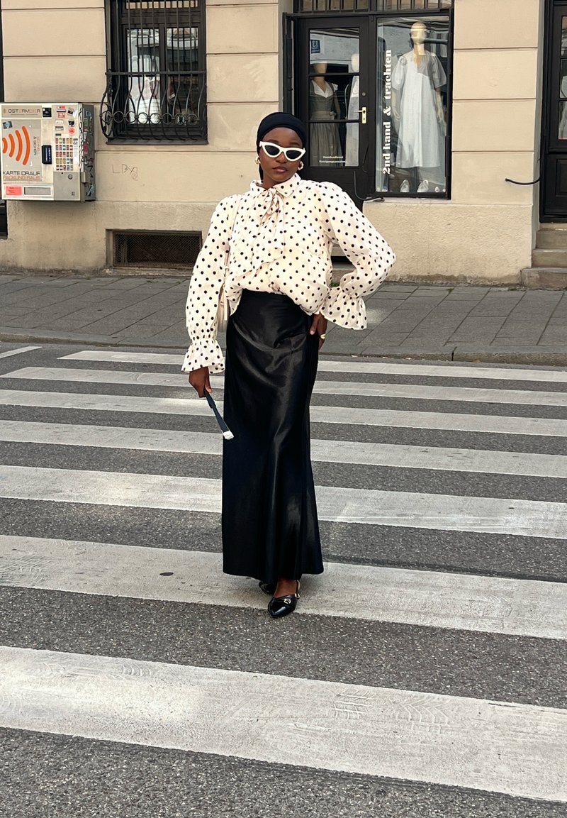 Femme portant un chemisier blanc à pois, une longue jupe noire et des lunettes de soleil blanches, debout avec assurance sur un passage pour piétons en ville.