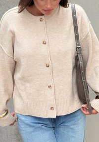 Beige kötött kardigán bordás aljjal és oversized viselettel, nagy gombokkal. Világos farmernadrággal és sötét keresztpántos táskával párosítva.