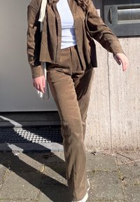 Persoană care merge în aer liber, purtând pantaloni și geacă maro din catifea, cu un top alb, purtând o geantă tote albă pe umăr.