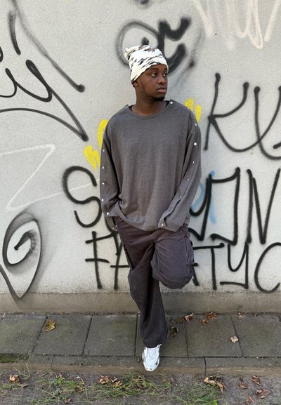 Top gris de manga larga con detalles de botones en las mangas, combinado con pantalones cargo oscuros y zapatillas deportivas blancas, frente a una pared graffiti.