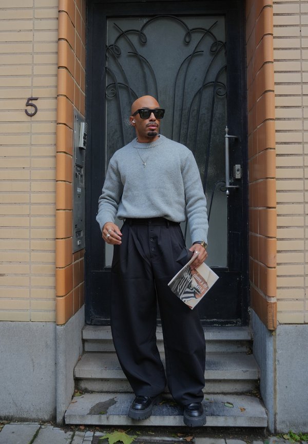 Un homme portant des lunettes de soleil, un pull gris et un pantalon noir ample se tient sur des marches devant une porte noire décorative, un magazine à la main.