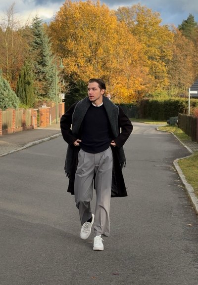 Persona vistiendo un abrigo negro con una bufanda gris, suéter azul marino y pantalones gris claro, caminando por una calle arbolada. Se ven zapatillas blancas.