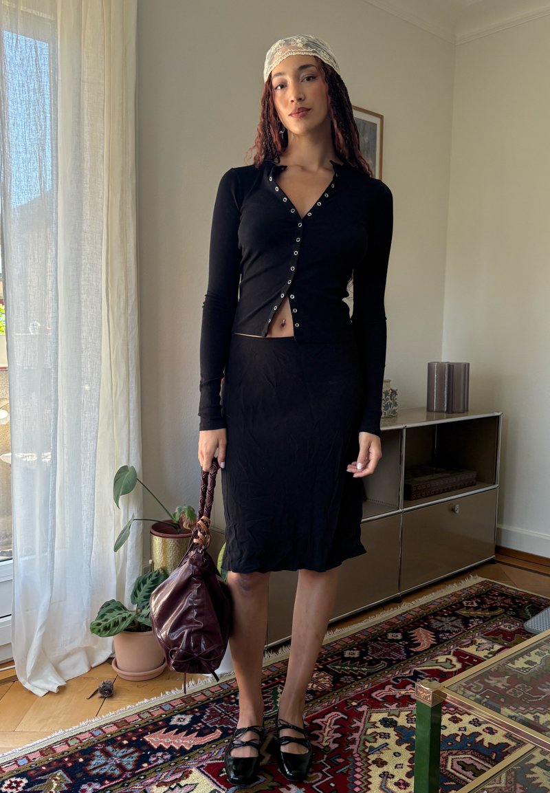 Femme debout à l'intérieur, portant un cardigan noir et une jupe, un foulard crème en dentelle sur la tête, tenant un sac en cuir marron, avec des plantes et des meubles en arrière-plan.