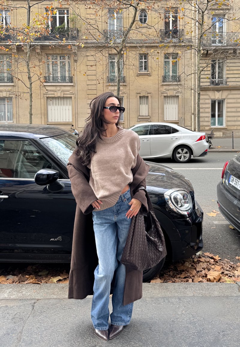 Beiger Strickpullover mit rundem Ausschnitt, kombiniert mit hellblauen ausgestellten Jeans. Brauner Mantel und strukturierte dunkle Handtasche. Schwarze Sonnenbrille vervollständigt das Outfit.