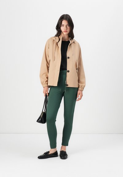 Chaqueta con capucha de color beige claro, top negro, pantalones ajustados verdes y zapatos negros. Presenta botones y un cordón ajustable. Incluye un bolso negro.