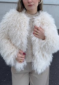 Γυναίκα που φοράει ένα χνουδωτό λευκό γούνινο μπουφάν πάνω από ένα μπεζ πουλόβερ και μπεζ παντελόνι, δείχνοντας πολλά χρυσά δαχτυλίδια και κόκκινο βερνίκι νυχιών.