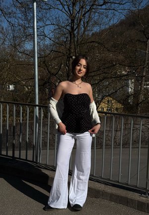 Joven con cabello oscuro de pie al aire libre cerca de una barandilla, vistiendo un top negro sin tirantes con lunares, pantalones blancos y una chaqueta blanca fuera de los hombros.