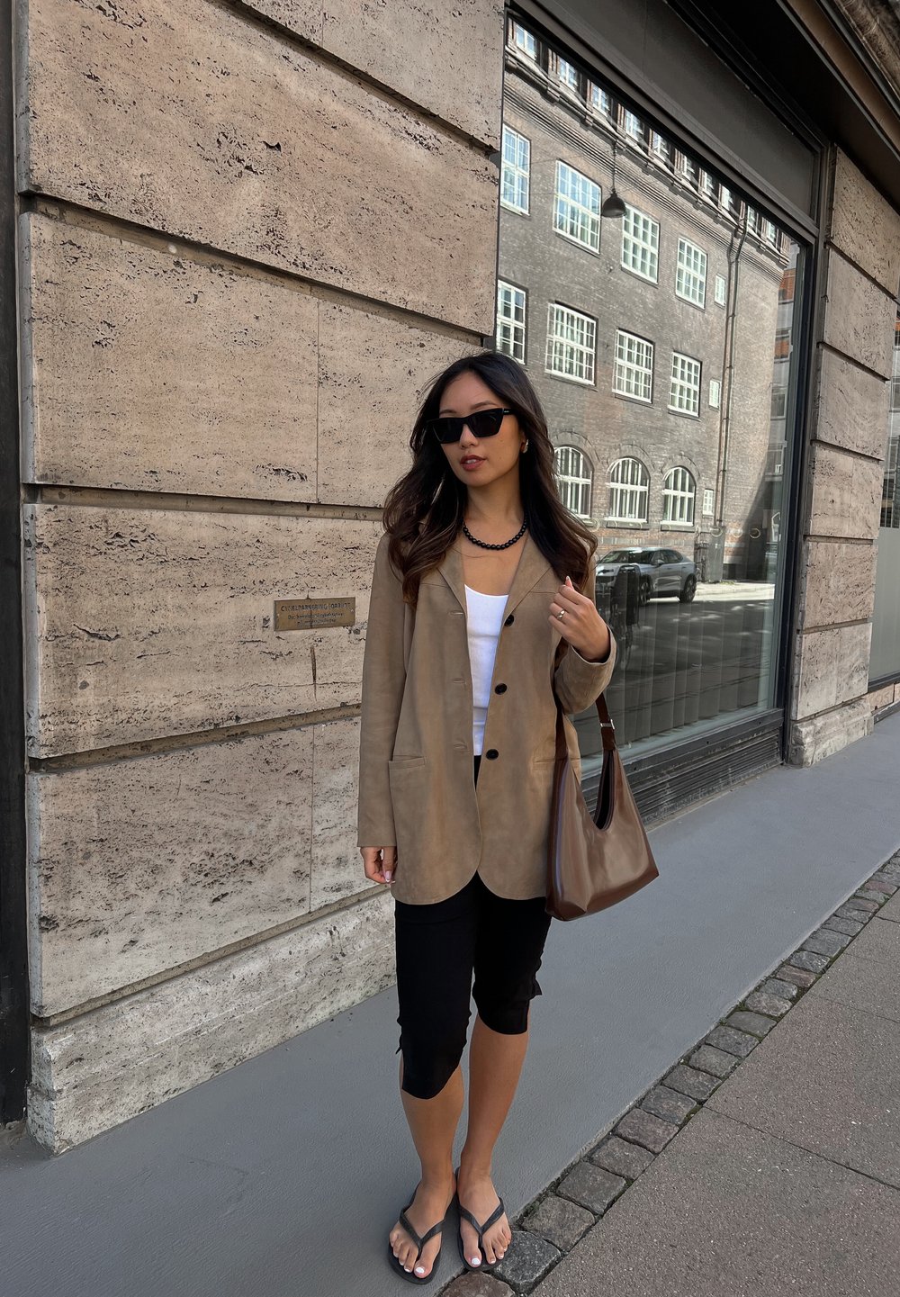 Καφέ δερμάτινο σακάκι με δύο μπροστινές τσέπες, λευκό αμάνικο μπλουζάκι, μαύρο κολάν και σαγιονάρες. Καφέ τσάντα ώμου και γυαλιά ηλίου.