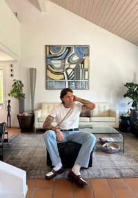 En tattoert person sitter på et svart ottoman, iført en hvit skjorte og lyse jeans. Et moderne kunstverk henger på veggen; en lyse farget sofa i bakgrunnen.