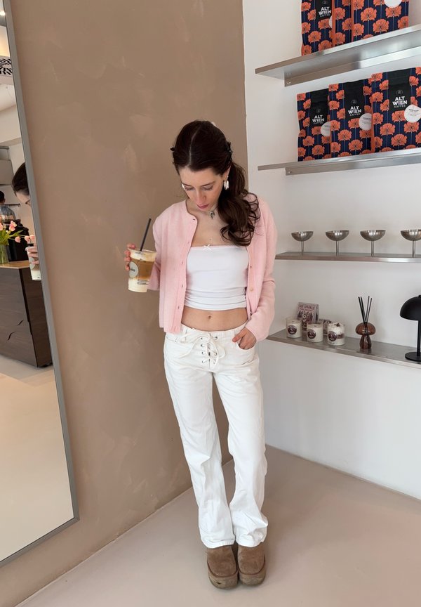 Ženska v roza jopici in belih hlačah, ki drži ledeno kavo, stoji ob regalih s paketi kave in svečami v minimalistični kavarni.