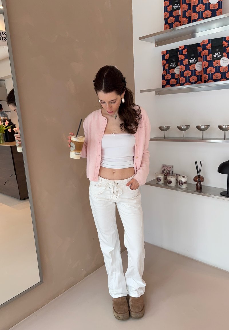 Nainen vaaleanpunaisessa neuletakissa ja valkoisissa housuissa, kädessään jääkahvi, seisoo hyllyjen vieressä, joissa on kahvipusseja ja kynttilöitä minimalistisessa kahvilassa.