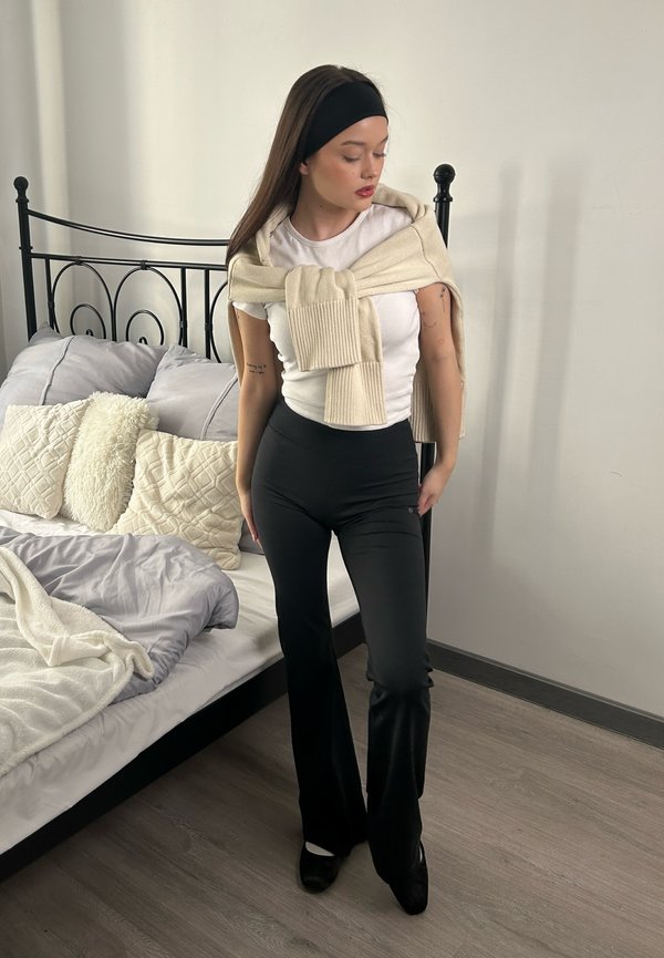 Vrouwelijke model draagt een wit T-shirt en zwarte flared broek, met een lichtbeige sweater over haar schouders. Zwarte haarband, binnenomgeving.