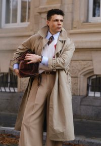 Trench coat beige sobre un suéter blanco con acentos rojos, pantalones crema y un bolso de cuero marrón. Presenta una camisa azul y una corbata estampada.