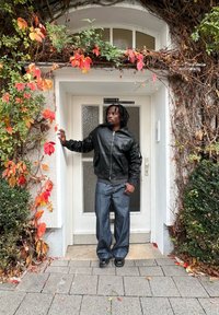 Melna ādas bomber jaka ar ribas aprocēm, izbalējušas zilas plataji džinsi un melni apavi pie durvīm, kas rotātas ar krāsainām rudens lapām.