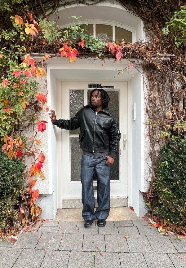 Chaqueta bomber de cuero negro con puños acanalados, vaqueros anchos de pierna en azul desgastado y zapatos negros frente a una puerta adornada con hojas otoñales de colores.