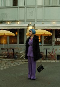 Costume ajusté violet avec un pantalon à jambes larges, manteau en fourrure noir et sac à main noir. Coiffure bob courte aux couleurs vives bleue et verte.