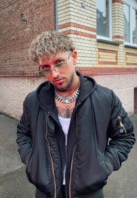 Svart bomberjakke med hette, glidelåslommer og ribbestrikkede mansjetter. Metallisk kjede halskjede og lyse solbriller. Bakgrunn av murvegg.