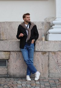 Mand i en sort dobbeltradet frakke med et brunt tørklæde, blå jeans og hvide sneakers, der læner sig op ad en stenvæg med en drink i nærheden.