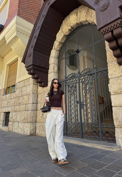 Modelo con una blusa marrón ajustada y pantalones anchos blancos sostiene un café. Lleva gafas de sol oscuras y zapatillas marrones, de pie junto a una pared de piedra.