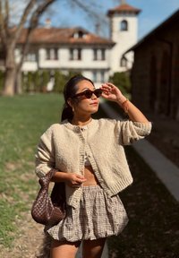 Cárdigan beige de punto con botones, top corto blanco, falda de cuadros marrón, bolsa de mimbre marrón y gafas de sol oscuro de carey en un entorno natural.