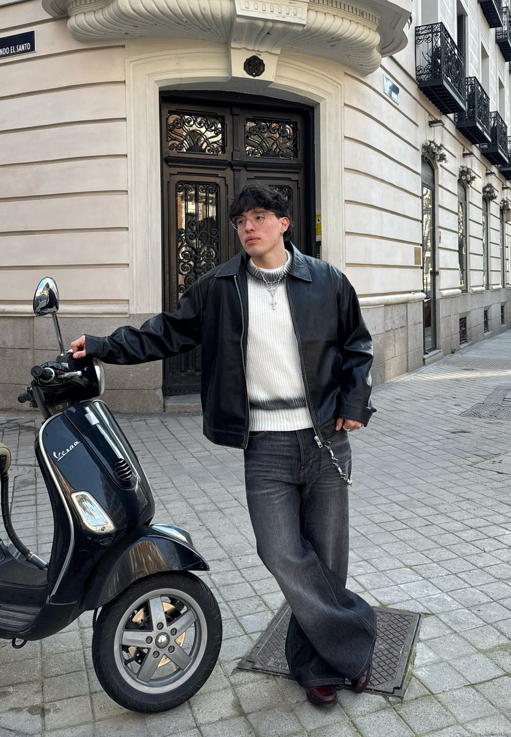 Trottinette noire avec un miroir chromé, garée sur des pavés; une personne en veste en cuir noir, pull blanc et jeans évasés s'appuie contre elle.