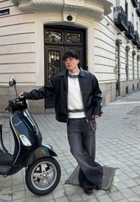 Trottinette noire avec un miroir chromé, garée sur des pavés; une personne en veste en cuir noir, pull blanc et jeans évasés s'appuie contre elle.