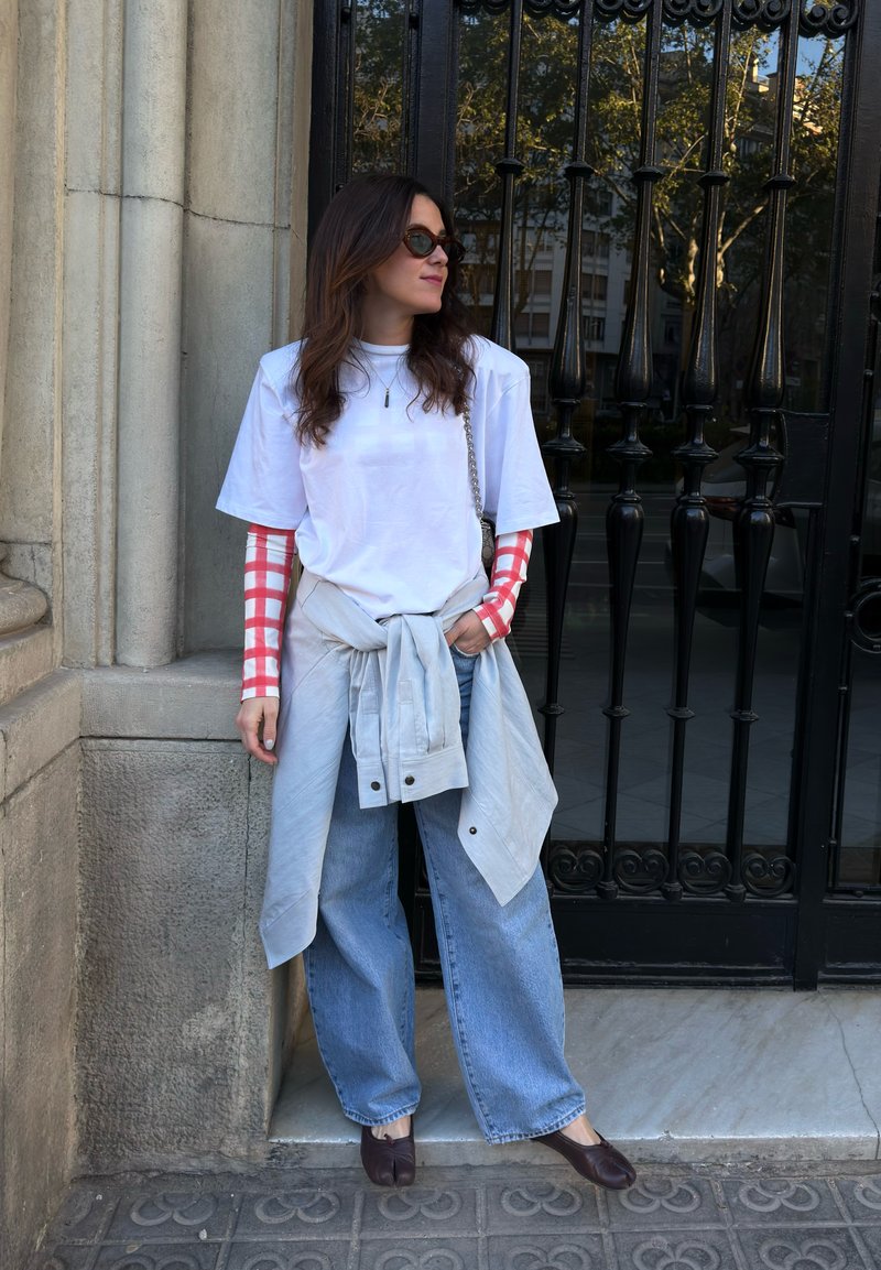 Woman with sunglasses wearing white T-shirt over red checkered long sleeves, blue wide jeans, tied jacket at waist, standing by black iron gate.