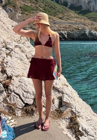 Bikini-Set in Burgunder mit strukturiertem Finish, kombiniert mit einem passenden Rock. Trägt einen Strohhut und lila Flip-Flops, während sie ein Buch hält.