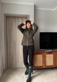 Γυναίκα σε εσωτερικό χώρο να ρυθμίζει σκούφο σκουρού χρώματος, φοράει χακί τζάκετ με φερμουάρ, μαύρο παντελόνι και σκούρα πράσινα λαστιχένια μποτάκια, στέκεται κοντά σε ξύλινη ντουλάπα και τηλεόραση.