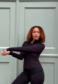 Schwarze Sportjacke mit Reißverschluss und langen Ärmeln, kombiniert mit schwarzen Leggings. Das Modell streckt die Arme vor einem hellgrünen Hintergrund aus.