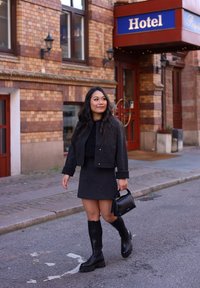 Veste noire cropped sur un haut texturé, assortie à une jupe ajustée. Bottes noires jusqu'aux genoux et petit sac à main noir. Mur en briques en arrière-plan.