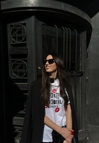 Czarny płaszcz na graficznej białej koszulce z odbitkami czerwonej szminki. Czarne okulary przeciwsłoneczne ozdabiają długie, ciemne włosy. W tle znajduje się ciemne drzwi.