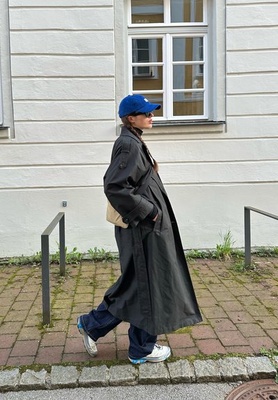 Abrigo largo negro con grandes solapas, detalles en beige, gorra azul, gafas de sol y zapatillas plateadas. El fondo presenta un camino y una pared de ladrillos.