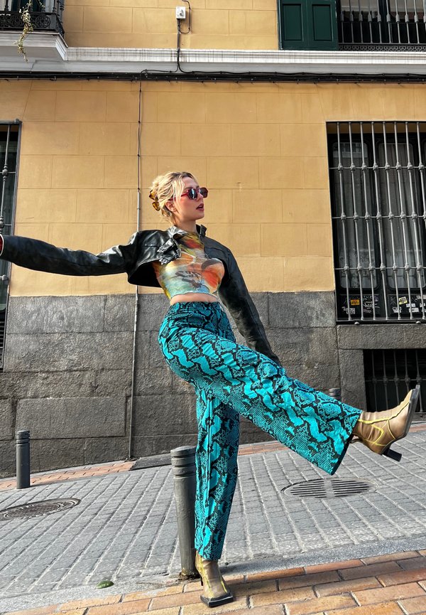 Zwarte leren cropped jas, kleurrijke graphic crop top, turkooise slangenprint wijde broek en metallic hoge hakken met straps.