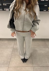 Grå croppet hettegenser med glidelås, kombinert med matchende høytlivsbukser, begge i mykt stoff. Sorte pumps med spiss tå fullfører antrekket.