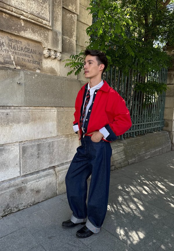 Jeune homme en veste rouge, chemise blanche, cravate rayée et pantalon en jean large, debout les mains dans les poches près d’un vieux mur en pierre et d’une clôture en fer.