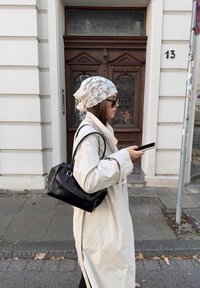 Světle barevný trenčkot, černá kabelka, krajkový šátek a sluneční brýle. Stojí vedle dřevěných dveří s jemnými detaily. Drží telefon.