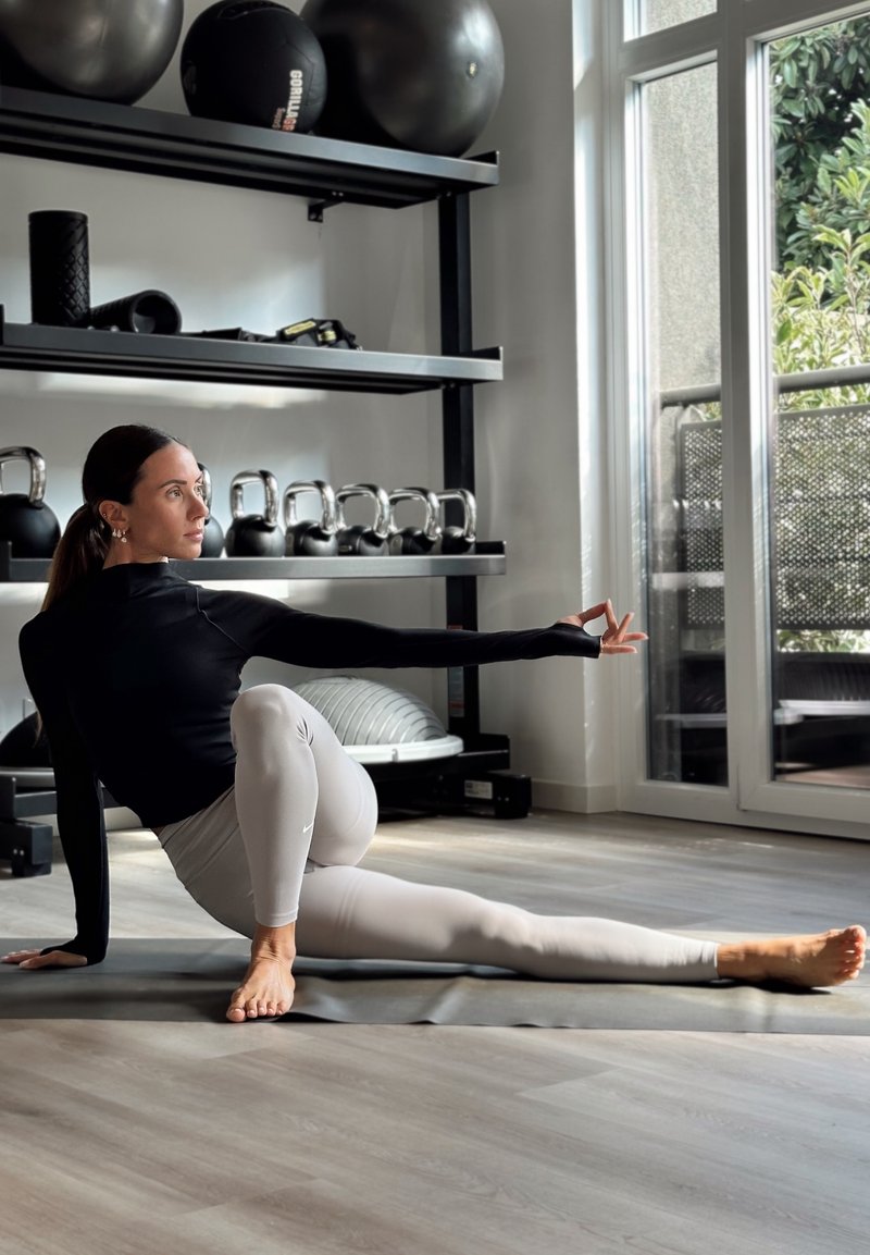 Aktīvās apģērbu komplekts ietver melnu garām piedurknēm tērpu un gaiši pelēkas legingus. Vide ir aprīkota ar fitnesa aprīkojumu un dabiska gaisma.