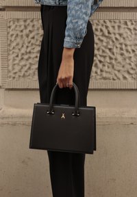 Fekete bőr kézitáska, amelynek szerkezeti téglalap alakja van, rövid fogantyúkkal és arany színű kiegészítőkkel. Egy modell viseli sötét nadrággal és farmerdzsekivel.