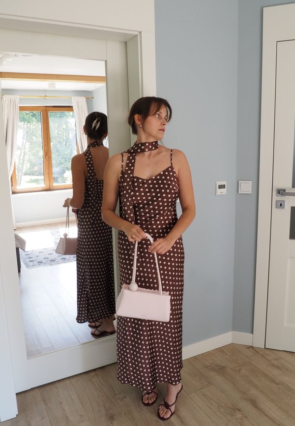 Femme en robe marron à pois tenant un sac à main rose clair, debout près d'un grand miroir dans une pièce lumineuse avec un sol en bois et une fenêtre.