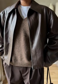 Άτομο που φοράει καφέ δερμάτινο μπουφάν πάνω από καφέ πλεκτή μπλούζα και λευκό πουκάμισο, με καφέ παντελόνι που ταιριάζει, στέκεται σε εσωτερικό χώρο με τα χέρια στις τσέπες.